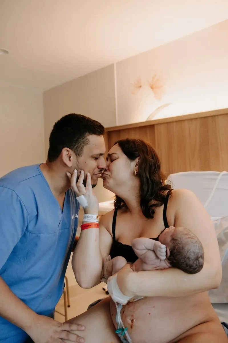 Pai beija a mãe com o bebê no colo no quarto do hospital
