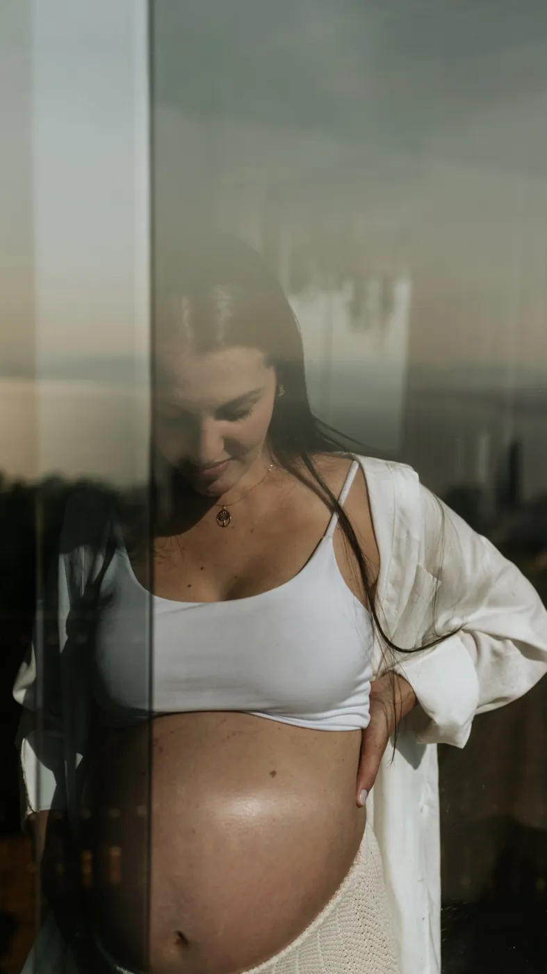 Gestante de top branco e camisa aberta junto à janela