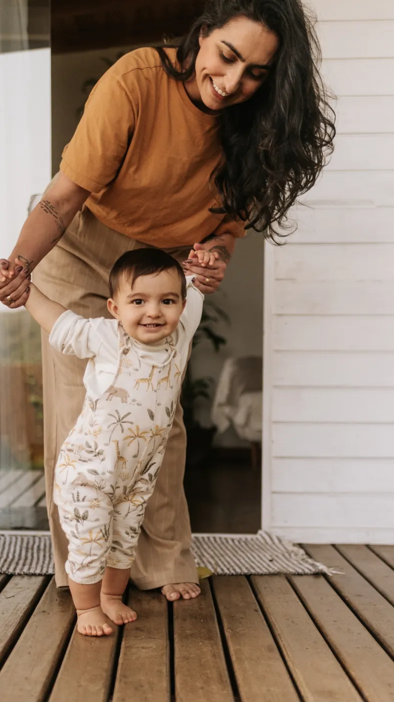 Mãe de camiseta mostarda segura as mãos do bebê de macacão estampado