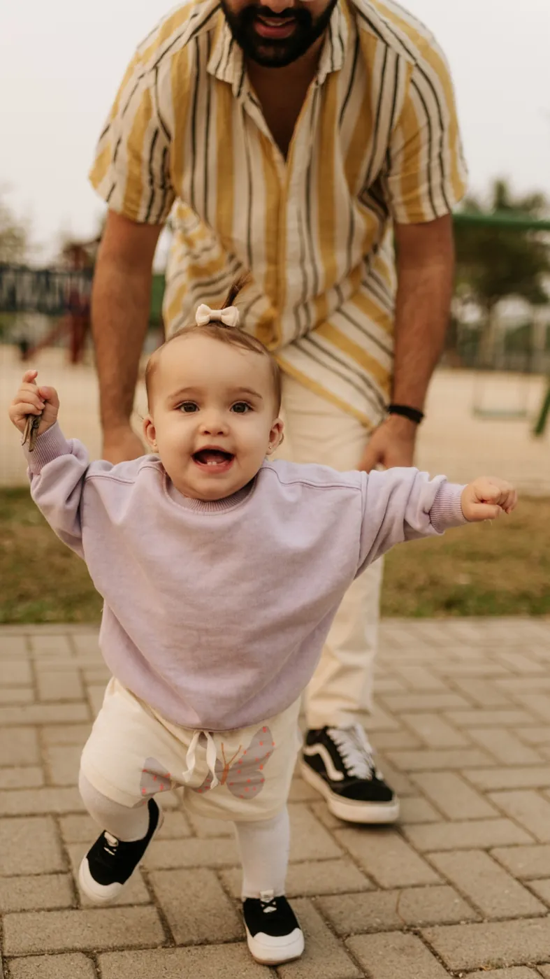 Bebê de blusa lilás dando passinhos com o pai de camisa listrada