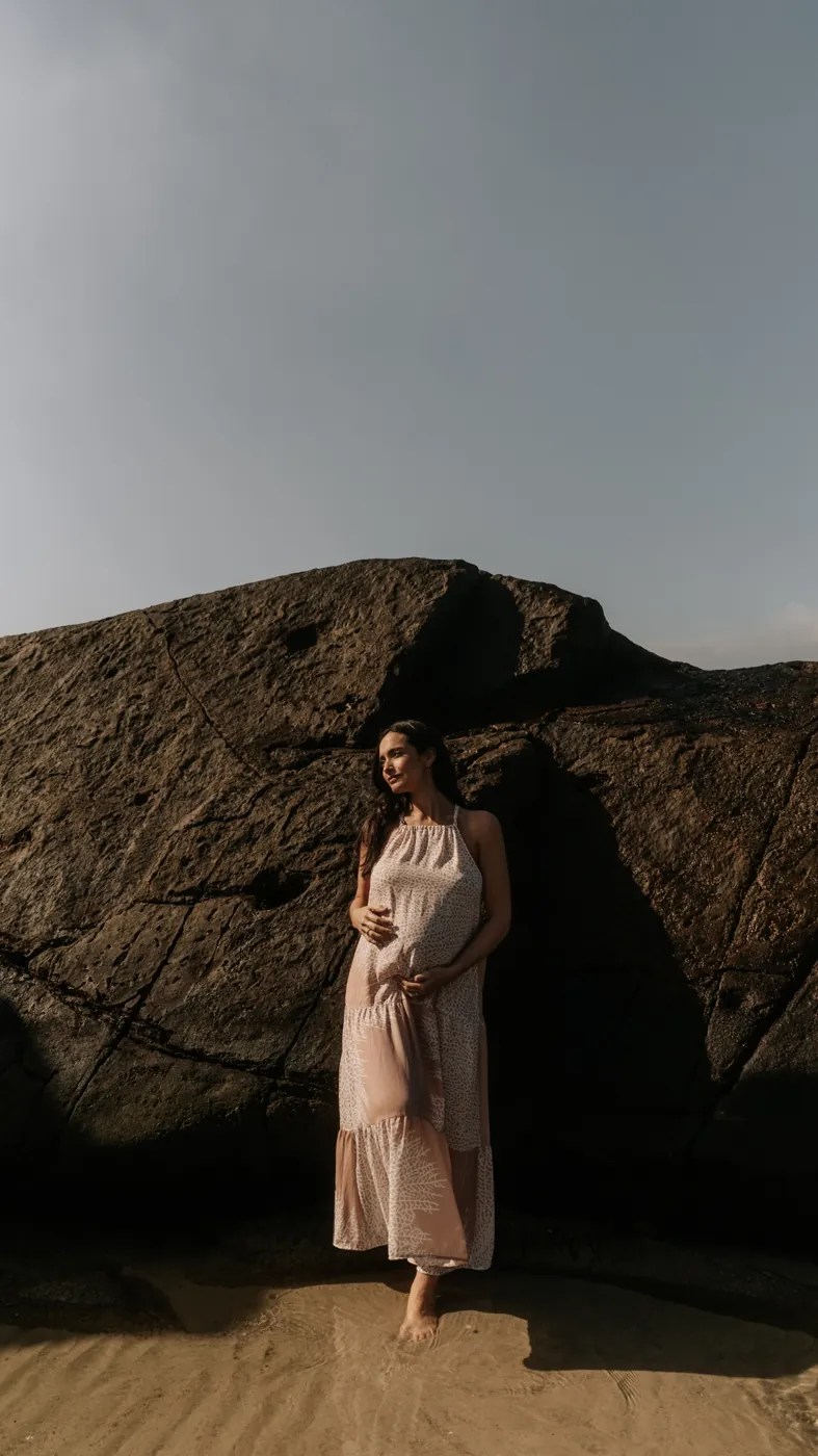 Gestante de vestido rosado junto a uma pedra na praia
