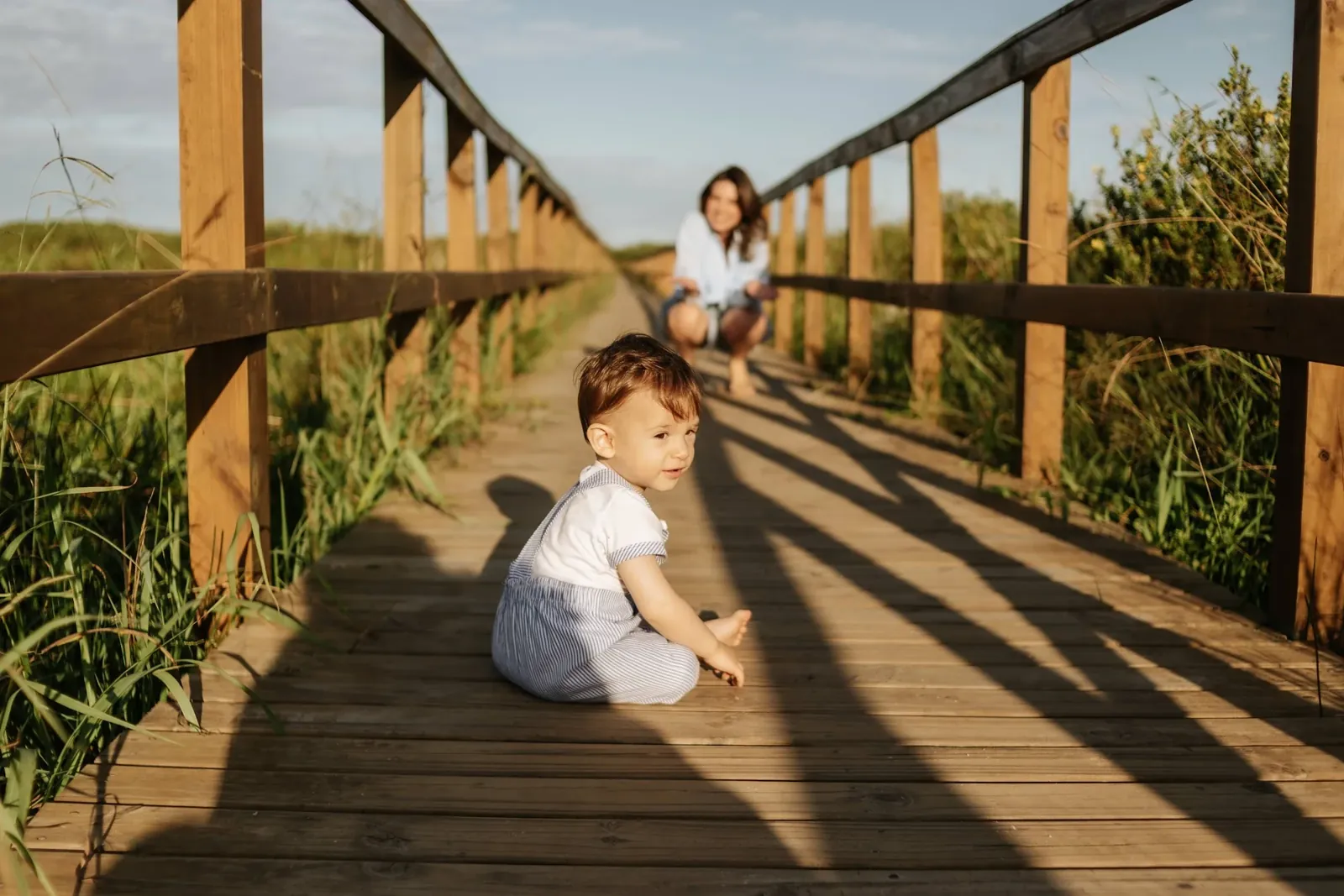 Bebê de nove meses sentado num deck de madeira, com a mãe ao fundo