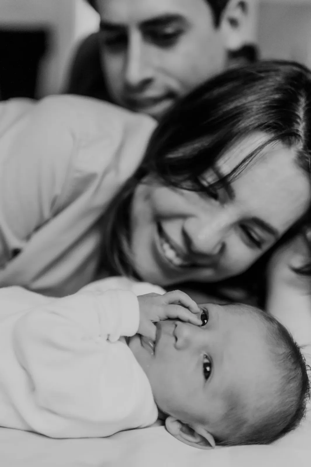 Pai e mãe sorriem deitados ao lado do recém-nascido, em preto e branco